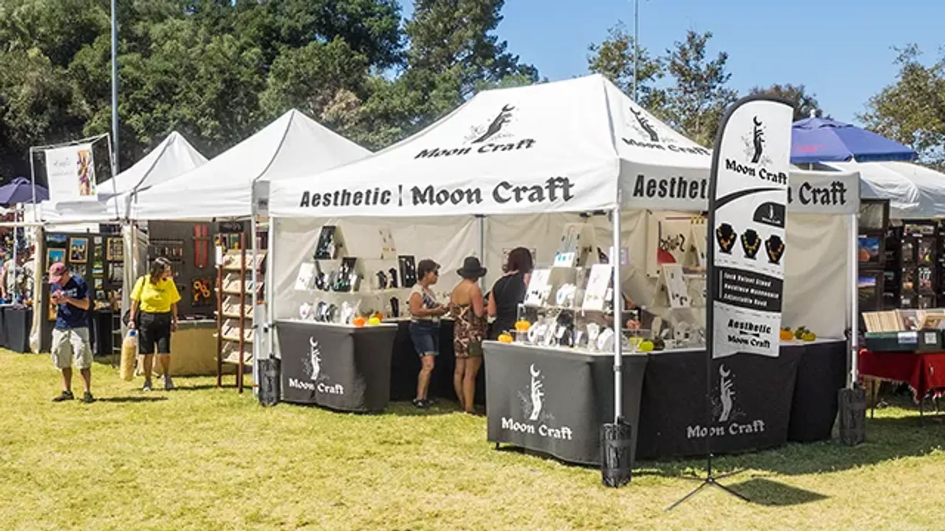 A custom white canopy tent with the "Moon Craft" logo set up at an outdoor craft fair, featuring a vendor displaying jewelry to browsing customers.
