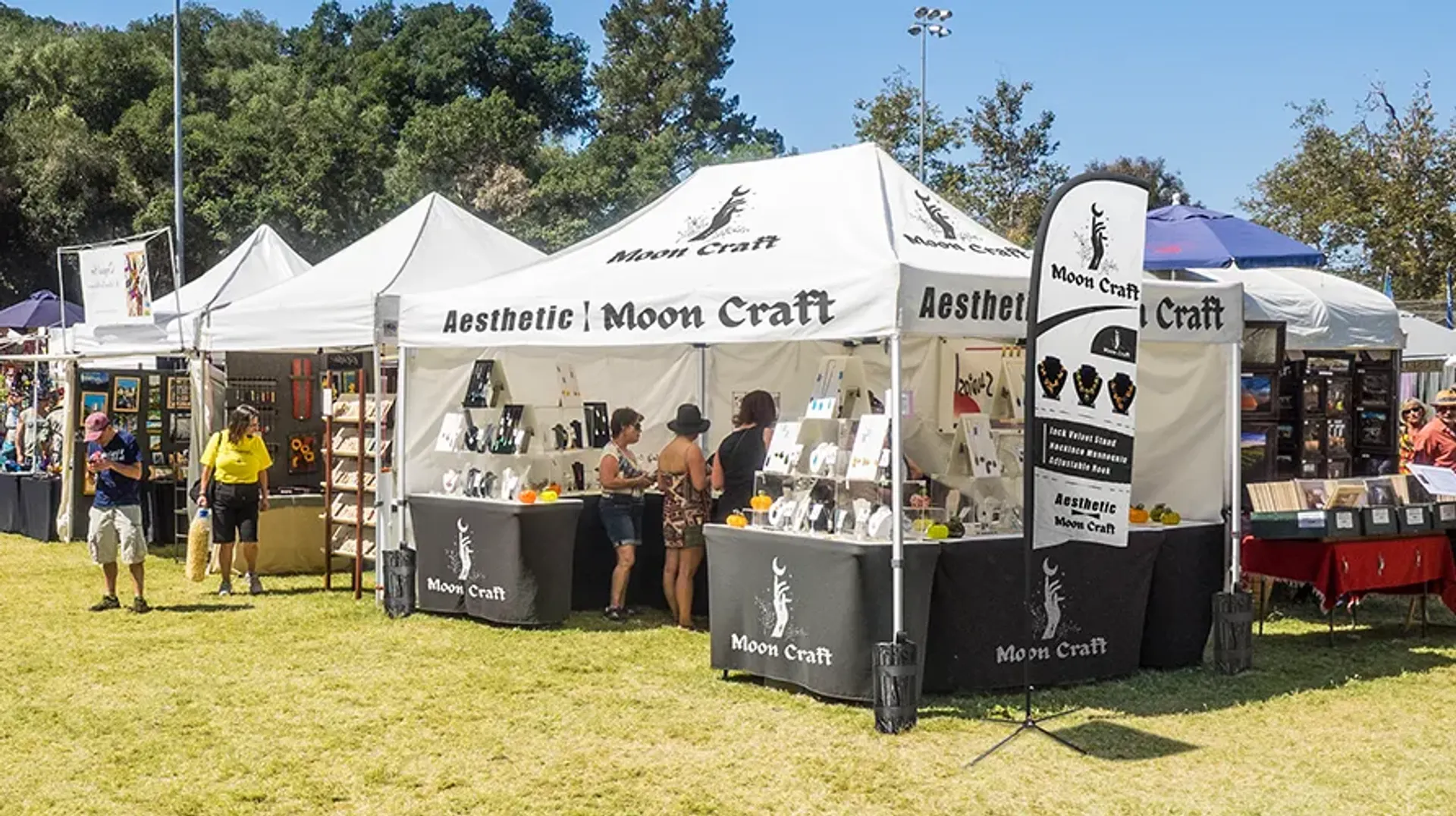 A custom white canopy tent with the "Moon Craft" logo set up at an outdoor craft fair, featuring a vendor displaying jewelry to browsing customers.
