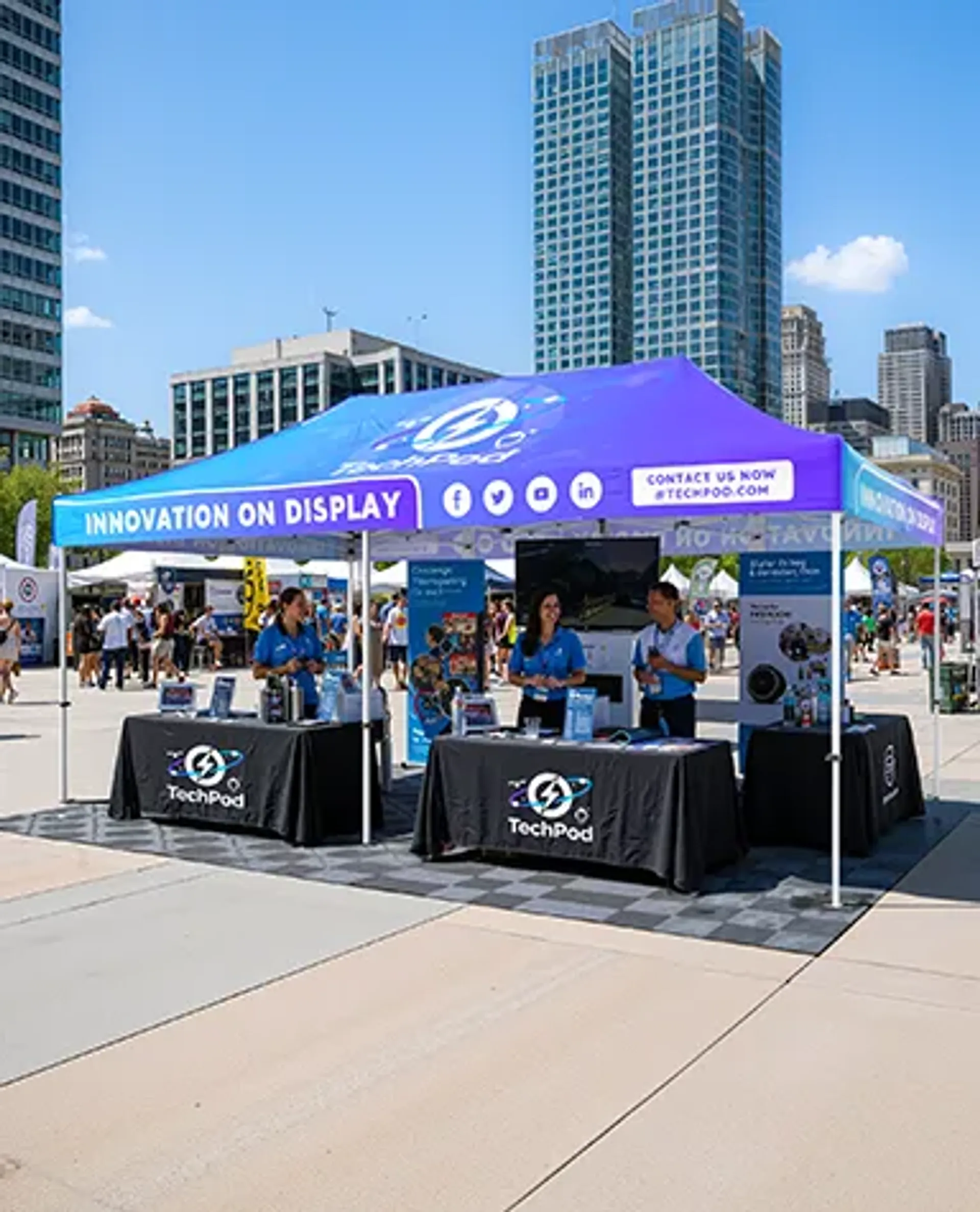 A commercial pop-up canopy with vibrant blue and purple custom graphics for "TechPod" located in an outdoor urban plaza, serving as a tech demonstration trade show booth with staff and display tables.
