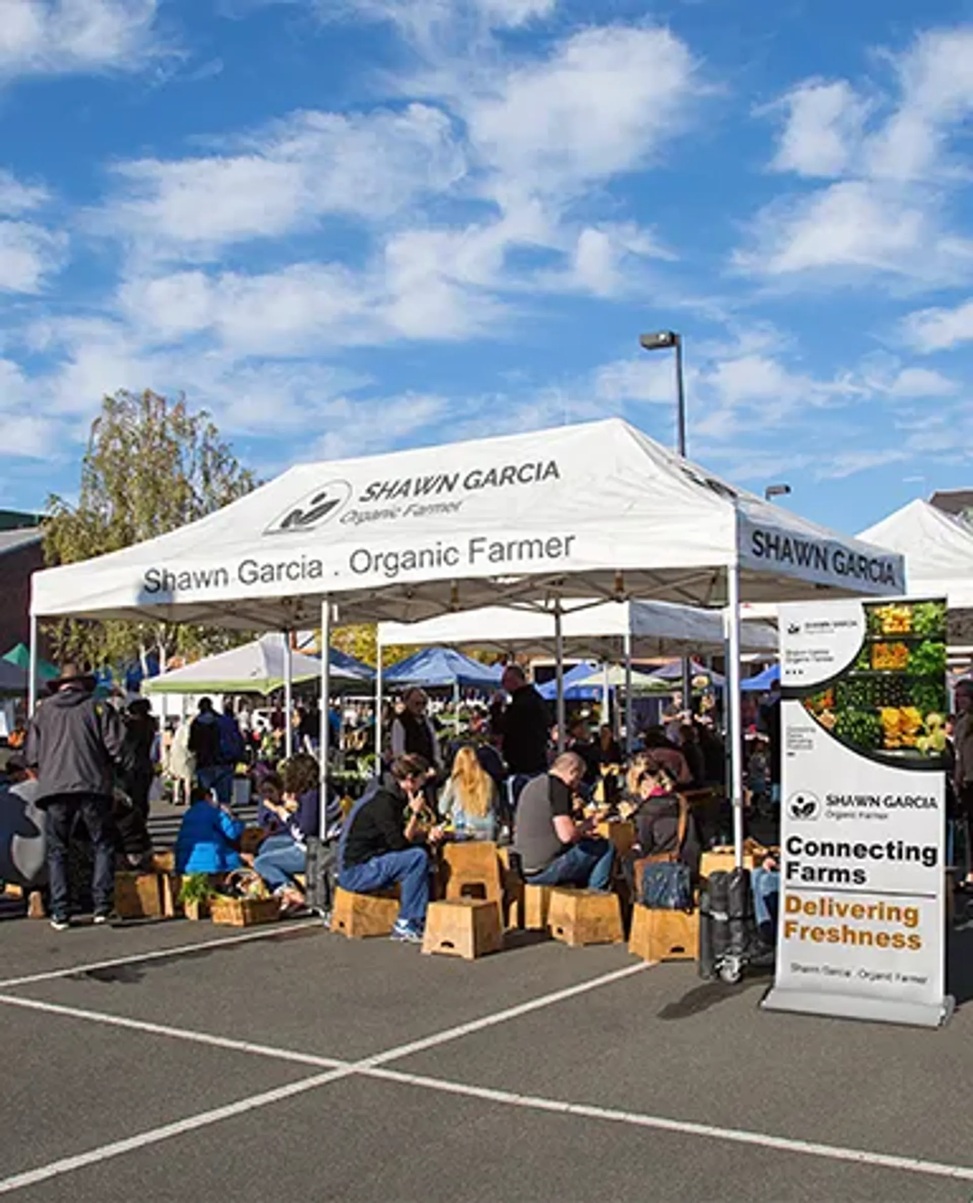 A white custom vendor tent for "Shawn Garcia Organic Farmer" at a bustling farmer's market, featuring branded text on the canopy top and an adjacent promotional banner stand.