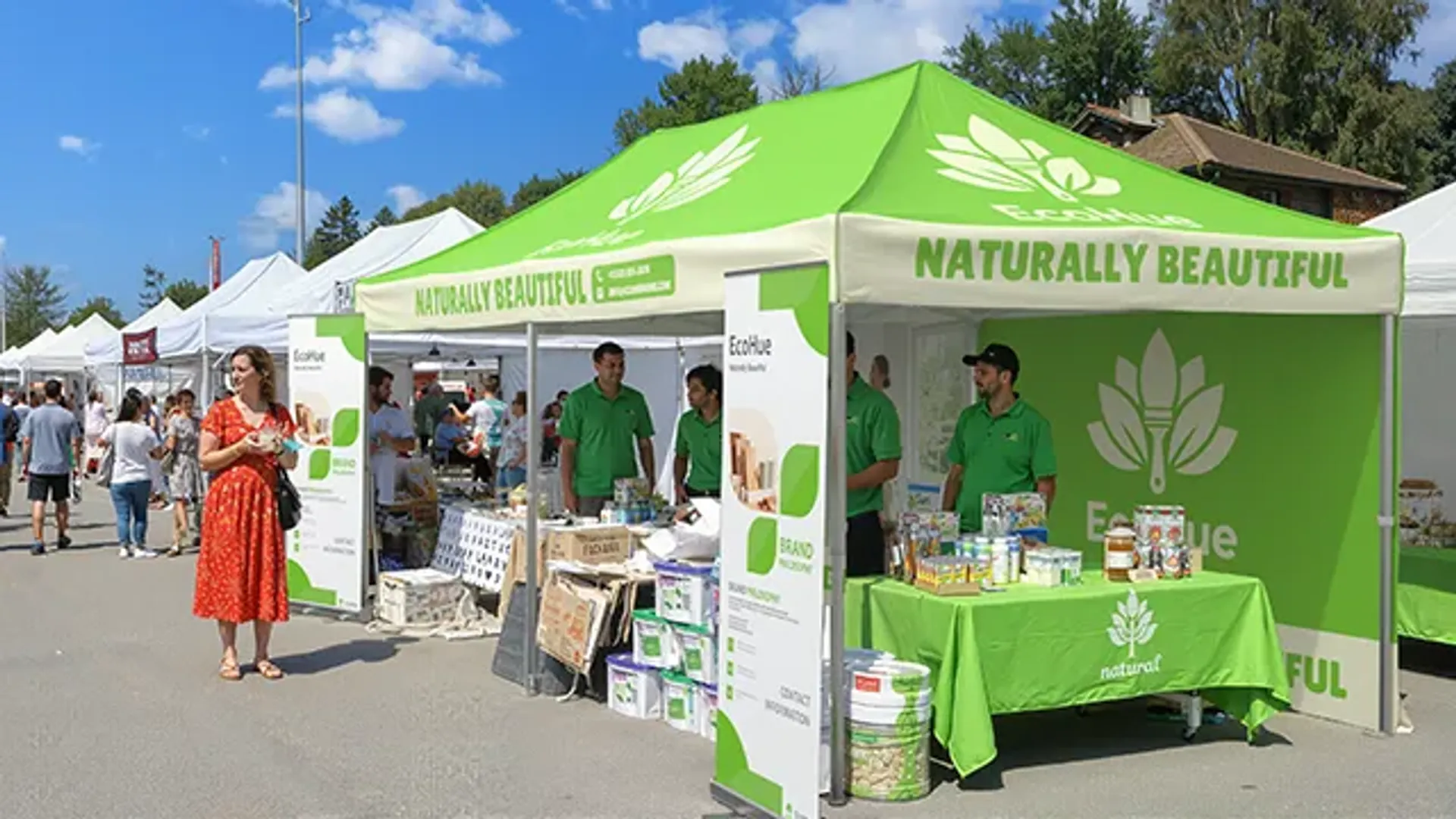 A custom printed green event canopy for the brand "EcoHue" at a sunny outdoor festival, showcasing natural products on a display table to eventgoers.