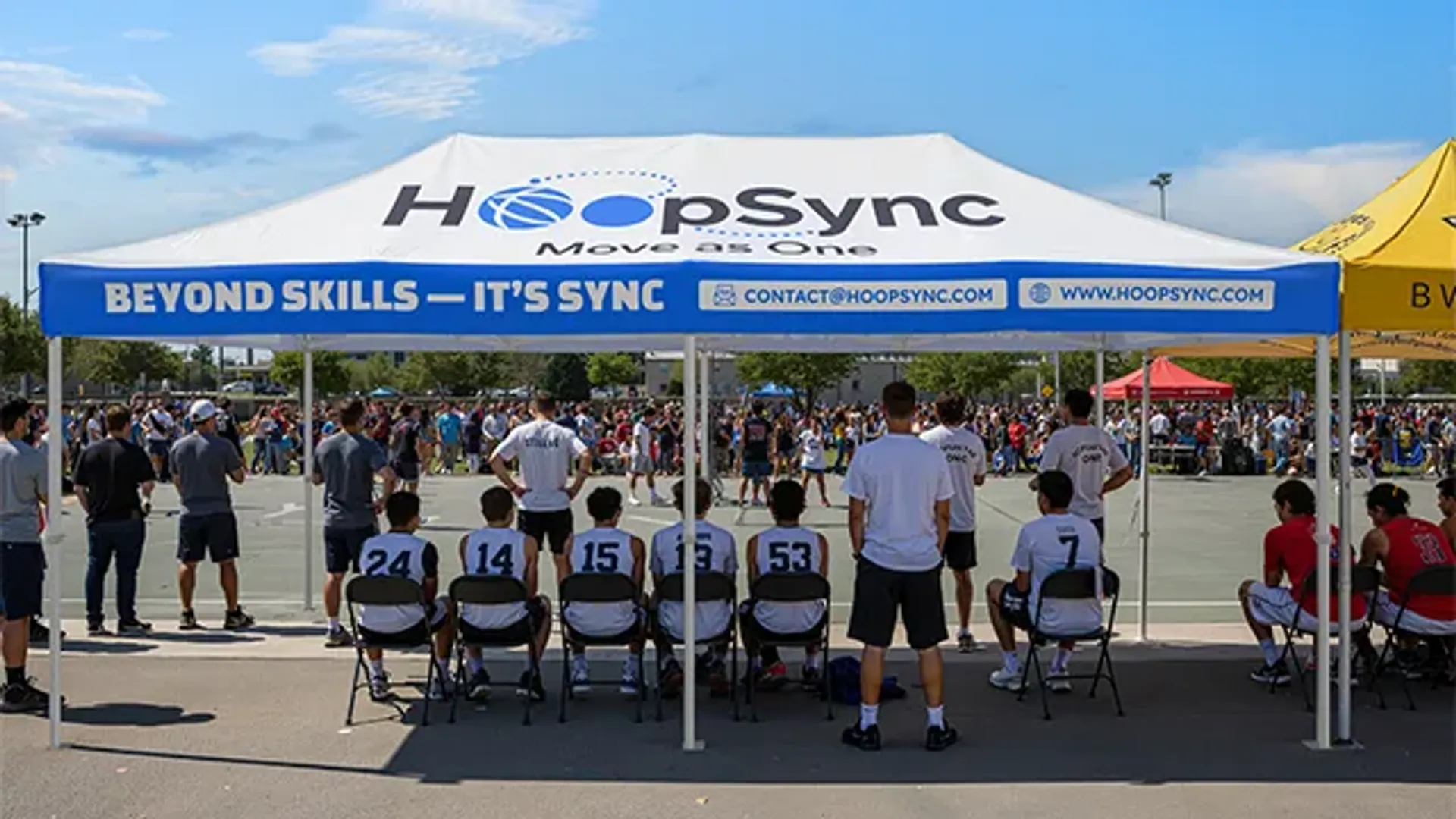 A large custom sports event tent with "HoopSync" branding, providing shade for basketball players sitting on the sidelines of an outdoor streetball tournament.
