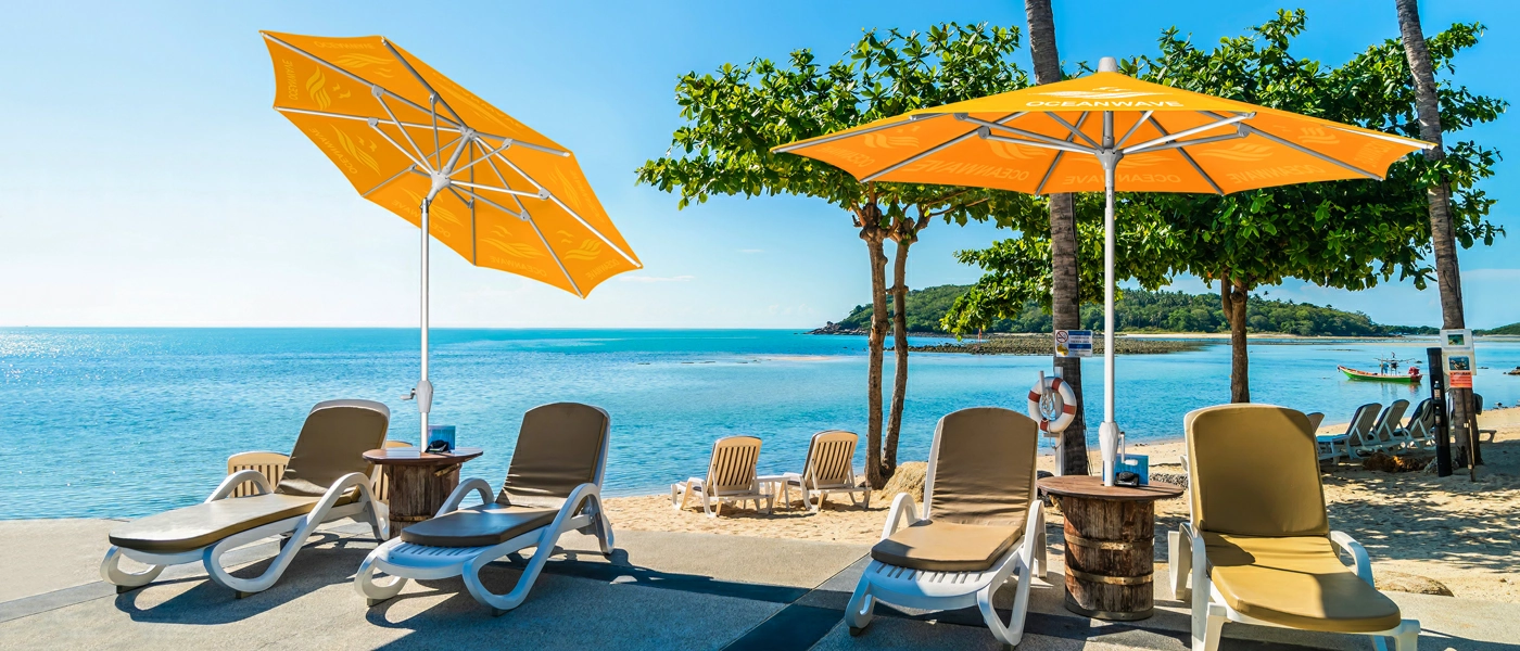 A row of lounge chairs and customiz patio umbrellas are placed on a serene beach. The tilting umbrellas are placed above the chairs to provide shade from the volume.