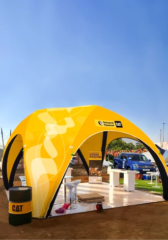 A large yellow custom inflatable tent, serves as an outdoor exhibition booth. A black and yellow barrel with a CAT logo stands next to the tent, and a blue pickup truck is visible in the background.